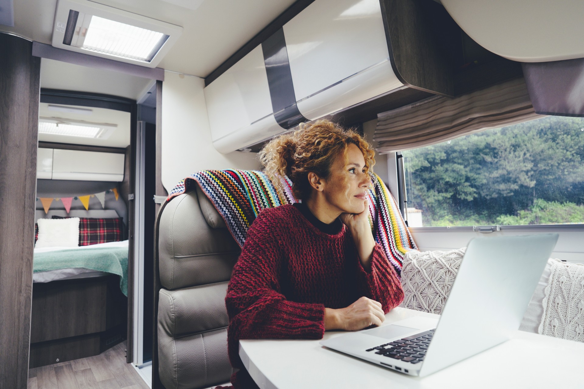 Adult woman use laptop computer inside a camper van recreational vehicle sitting at the table with bedroom in background and nature park outside the windos. Concept of travel and remote worker female people